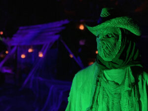 Halloween 2020 - Halloween actor in scarecrow costume under green lighting with creepy barn on path with candles and under purple lighting